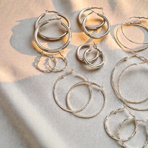 Könnte beinhalten: Eine Auswahl an silbernen Creolen in verschiedenen Größen und Designs. Die Ohrringe umfassen klassische glatte Creolen, strukturierte Creolen und kleinere, filigranere Stile. Der Schmuck ist auf einem weichen, hellfarbigen Stoff angeordnet, mit Schatten.