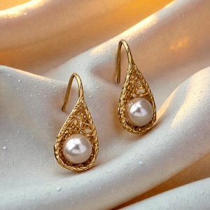 Può includere: Un paio di orecchini pendenti color oro. Ogni orecchino presenta una cornice a forma di goccia intrecciata che racchiude una grande perla bianca e rotonda. Gli orecchini pendono da un gancio curvo. Lo sfondo è un morbido tessuto bianco.