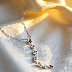Può includere: Una collana d'argento con una serie di pietre preziose colorate di varie forme. Le pietre preziose includono forme rettangolari, ovali e rotonde in tonalità di rosso, verde e marrone. La collana è esposta su un tessuto bianco e oro.