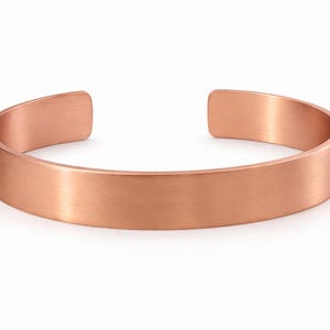 Op de afbeelding: Een koperen manchetarmband met een geborstelde afwerking. Het open ontwerp heeft een platte, rechthoekige band die zachtjes buigt. De armband heeft een warme, roodbruine kleur en lijkt van metaal te zijn.
