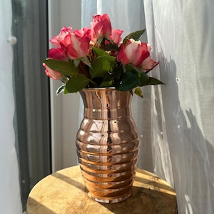 Könnte beinhalten: Eine kupferfarbene Vase mit geripptem Design hält einen Strauß rosa und weißer Rosen mit grünen Blättern. Die Vase steht auf einer Holzoberfläche, mit einem durchsichtigen weißen Vorhang im Hintergrund.