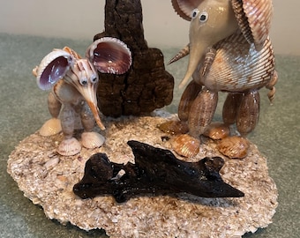 May include: Two whimsical seashell elephant sculptures with cockle shell ears and trunks, standing on a base of shells and sand. A piece of dark driftwood is behind them. The sculptures are tan, brown, and white.