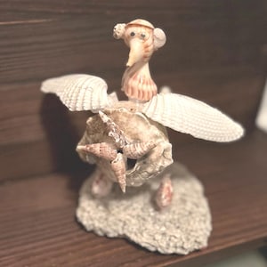 May include: A whimsical seashell sculpture of a bird with outstretched wings. The bird is made of various seashells, including a large shell for the body, smaller shells for the wings, and a spiral shell for the head. The bird is perched on a base of coral.