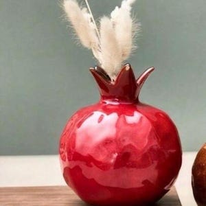 May include: A glossy, bright red ceramic vase shaped like a pomegranate with a textured, uneven surface. The top of the vase features a crown-like opening holding several fluffy, cream-colored dried grass stems that extend upwards.