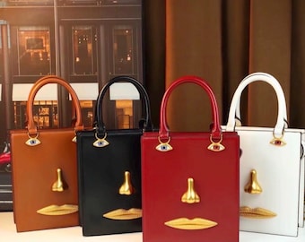 Puede incluir: Cuatro bolsos estructurados en color marrón claro, negro, rojo y blanco. Cada bolso presenta un adorno dorado de nariz y labios en la parte frontal, con dos dijes en forma de ojo sujetos a la base de las asas, creando un diseño facial estilizado.