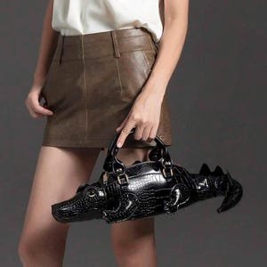 May include: A black handbag shaped like a crocodile with a textured, embossed surface that mimics reptile scales. The bag features a pointed snout, gold-tone hardware accents on the handles, and a structured body with a tail extending from the back.