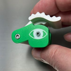 May include: A 3D printed mechanical fidget toy featuring a bright green body with a white eye graphic and a white scalloped top piece. The device includes two small metallic hex screws visible on the green surface, providing a tactile mechanism.