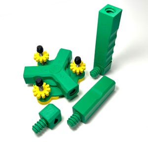 May include: Green plastic construction toy set with various components, including a cross-shaped piece with yellow and black accents, a handle, and several screw-like parts. The pieces are arranged on a white surface.