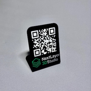 Könnte beinhalten: Ein schwarzer QR-Code-Ständer mit weißem QR-Code und dem Text "NextLayer 3D Studio" in Weiß und Grün. Der Ständer hat eine rechteckige Form mit abgerundeten Ecken und einem kleinen Sockel. Der Hintergrund ist grau.
