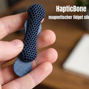 Könnte beinhalten: Ein dunkelblauer HapticBone magnetischer Fidget-Slider mit einer texturierten Wabenstruktur auf der Oberseite und einer glatten, abgerundeten Basis. Der Text HapticBone magnetischer fidget slider ist in Weiß rechts im Bild aufgedruckt.