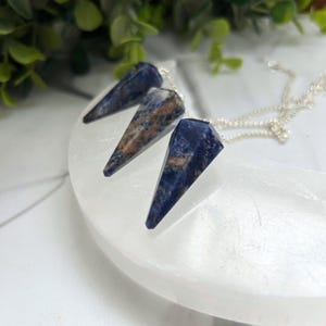 May include: Three blue sodalite crystal pendulums with silver chains rest on a white, curved surface. The pendulums have a pointed shape and feature brown and white marbling. Green foliage is visible in the background.