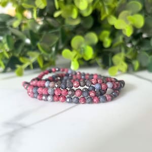 May include: A multi-strand bracelet made of small, faceted beads in red, blue, and grey. The beads are strung together, creating a flexible, coiled design. The bracelet is shown on a white surface with green foliage in the background.