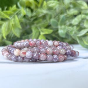 May include: A strand of round, faceted beads in shades of pink, purple, and white. The beads are arranged in a spiral, with a blurred green leafy background. The beads appear to be made of a natural stone.