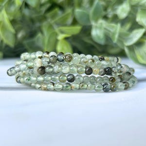 May include: A bracelet made of small, round, green prehnite beads. The beads have a translucent quality with darker green and brown inclusions. The bracelet is coiled and rests on a white surface, with blurred green foliage in the background.
