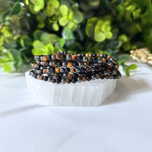 Pode incluir: Uma pilha de pulseiras de contas com uma mistura de contas pretas, castanhas e castanho-dourado. As pulseiras estão sobre uma superfície branca e texturizada, com folhagem verde ao fundo. As contas parecem pequenas e redondas.