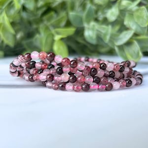 May include: A stack of beaded bracelets featuring a mix of pink, red, and black beads. The bracelets are made of small, round beads and are arranged in a spiral. The background is blurred, with green foliage.