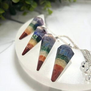 May include: Four gemstone pendulums with a rainbow color scheme, each suspended from a silver chain. The pendulums are arranged on a white, crescent-shaped surface, with green foliage in the background. The gemstones are faceted and have a pointed tip.