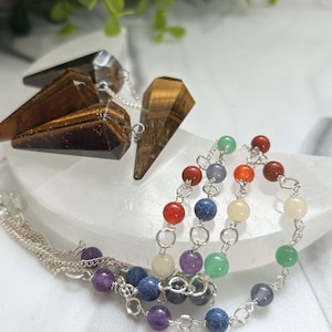 May include: Three tiger's eye crystal pendulums with silver chains are on a white surface. A beaded chakra necklace with colourful gemstones is also visible. The gemstones include red, orange, yellow, green, blue, and purple.