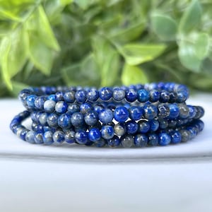 May include: Several strands of blue lapis lazuli gemstone bracelets. The beads are a deep blue color with flecks of gray and gold. The bracelets are stacked on top of each other, with a blurred green background.