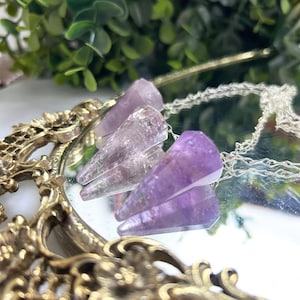 May include: Three amethyst crystal pendants on silver chains, displayed on a decorative mirror. The crystals are a deep purple color with a pointed shape. The mirror has a gold ornate frame, and greenery is visible in the background.