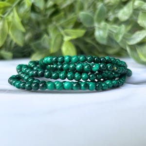 May include: A collection of dark green malachite beaded bracelets. The bracelets are coiled on a white surface, with a blurred green leafy background. The beads are small and round, showcasing the natural patterns of the stone.