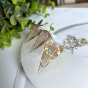 May include: Three clear quartz crystal pendulums with silver chains. The crystals are faceted into a pointed, triangular shape. The pendulums are displayed on a white, curved surface, with green foliage in the background.