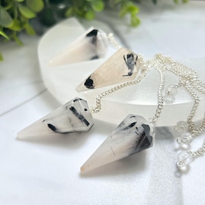 May include: Four pendulums with silver chains and clear quartz beads. The pendulums are white with black inclusions and a pointed shape. They are displayed on a white surface with green foliage in the background.