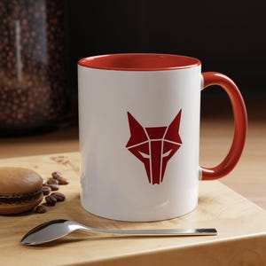 May include: White ceramic mug with a red interior and handle. The mug features a geometric red fox head design on the front. A spoon, a macaron, and coffee beans are on a wooden surface next to the mug.