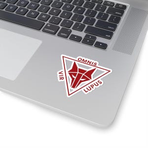 Może przedstawiać: Naklejka z czerwonym wzorem głowy wilka w białym trójkącie, ze słowami "OMNIS VIR LUPUS" na czerwono. Naklejka jest umieszczona na laptopie.