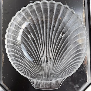 May include: Clear glass seashell-shaped serving dish. The dish has a scalloped edge and a ribbed, fan-like design. The dish is on a black tray.