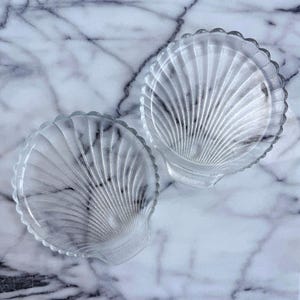 May include: Two clear glass seashell-shaped dishes. Each dish has a scalloped edge and a ribbed interior design. The dishes are displayed on a white and gray marble surface, creating a coastal or nautical theme.