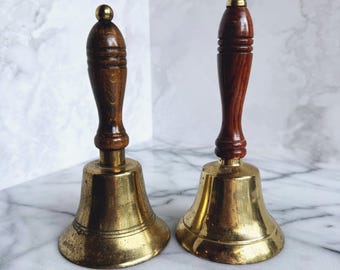 May include: Two antique brass bells with wooden handles. The bells have a classic design with a flared base and a rounded top. The handles are made of dark wood and are topped with a small brass sphere. The bells are likely used for calling or signaling.