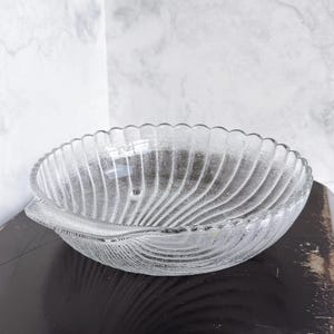 May include: Clear glass serving bowl with a scalloped rim and a textured, ridged design. The bowl has a leaf-like shape with a small handle on one side. The bowl is sitting on a dark surface.