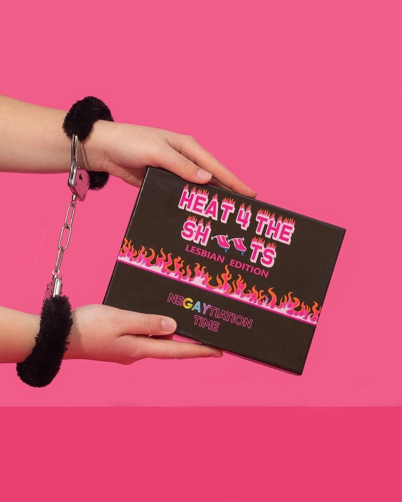 Op de afbeelding: Een zwarte doos met de tekst "HEAT 4 THE SHOTS LESBIAN EDITION" en "NEGAYTIATION TIME" in roze en witte letters. De doos is omringd door vlammen. Een persoon houdt de doos vast met zwarte, harige handboeien om de pols.