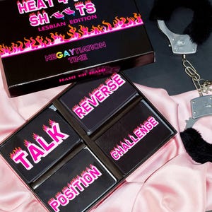 Op de afbeelding: Een zwarte doos met de tekst "HEAT 4 THE SH*TS LESBIAN EDITION" en "NEGAYTIATION TIME" op de deksel. De doos bevat vier zwarte kaarten met de woorden "TALK", "POSITION", "REVERSE" en "CHALLENGE" in roze tekst met vlammen. Er is ook een paar zwarte, harige handboeien te zien.