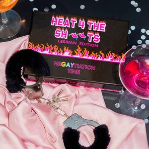 Op de afbeelding: Een zwarte doos met de tekst "HEAT 4 THE SHOTS LESBIAN EDITION NEGATIATION TIME" erop. De doos staat op een zwart oppervlak met zilveren confetti. Op een roze satijnen oppervlak liggen twee zwarte, harige handboeien met zilveren metalen sluitingen en sleutels.