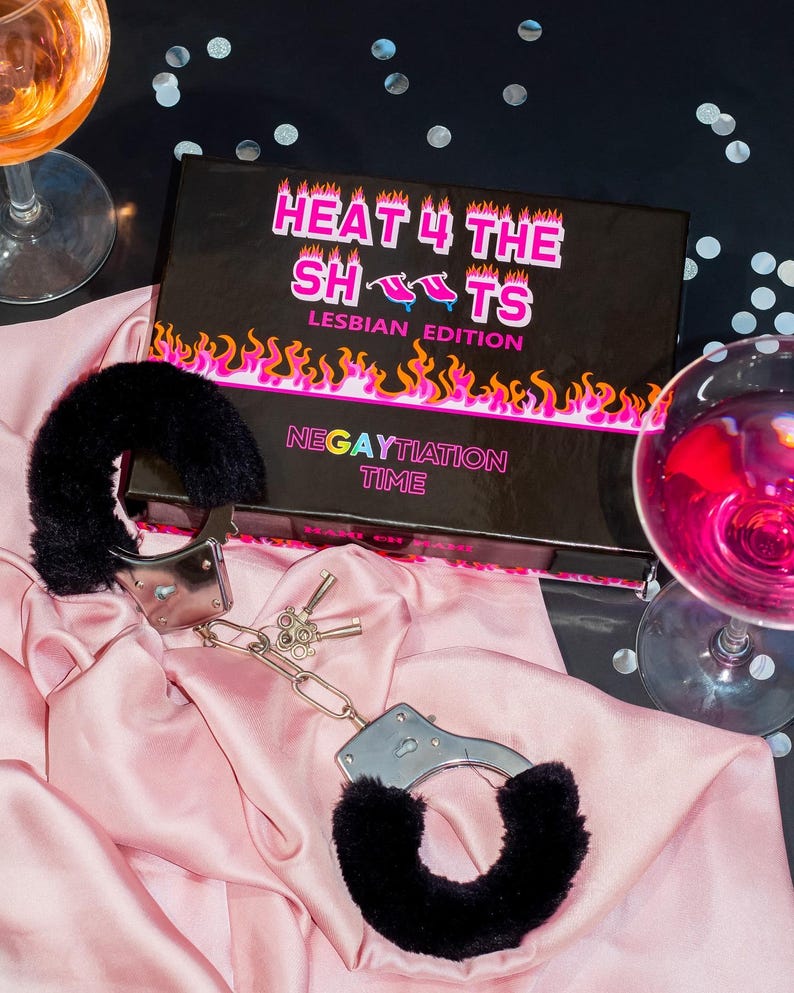 Op de afbeelding: Een zwarte doos met de tekst "HEAT 4 THE SHOTS LESBIAN EDITION NEGATIATION TIME" erop. De doos staat op een zwart oppervlak met zilveren confetti. Op een roze satijnen oppervlak liggen twee zwarte, harige handboeien met zilveren metalen sluitingen en sleutels.