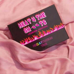 Op de afbeelding: Een zwarte doos met de tekst "HEAT 4 THE SHOTS LESBIAN EDITION" in roze en witte letters. De doos heeft een vlammenontwerp met de tekst "NEGAYTIATION TIME" in regenboogkleuren.