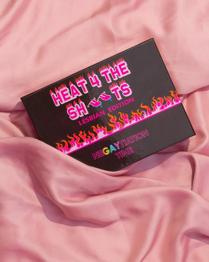 Op de afbeelding: Een zwarte doos met de tekst "HEAT 4 THE SHOTS LESBIAN EDITION" in roze en witte letters. De doos heeft een vlammenontwerp met de tekst "NEGAYTIATION TIME" in regenboogkleuren.