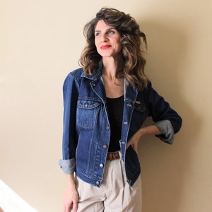 Puede incluir: Una mujer con una chaqueta vaquera azul sobre un top negro y pantalones beige con un cinturón marrón. Está de pie frente a una pared de color beige claro.