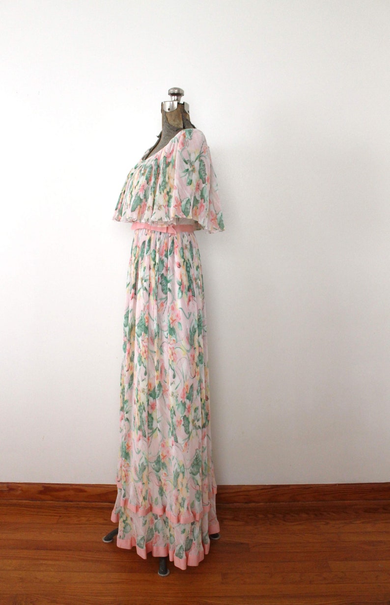 May include: A long, white maxi dress with a pink floral print and a ruffled neckline. The dress has a pink sash at the waist and a tiered skirt.