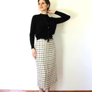 May include: A woman wearing a black cardigan and a white and black plaid skirt. She is standing in front of a white wall and has her hair pulled back.