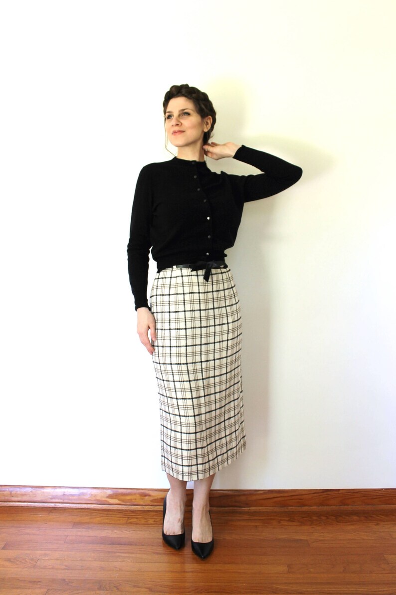 May include: A woman wearing a black cardigan and a white and black plaid skirt. She is standing in front of a white wall and has her hair pulled back.