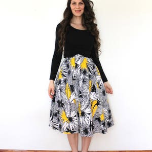 May include: A woman wearing a black long-sleeved shirt and a white and yellow floral skirt. She is standing on a wooden floor in front of a white wall.