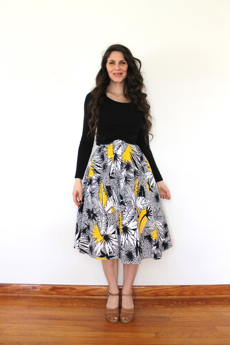 May include: A woman wearing a black long-sleeved shirt and a white and yellow floral skirt. She is standing on a wooden floor in front of a white wall.