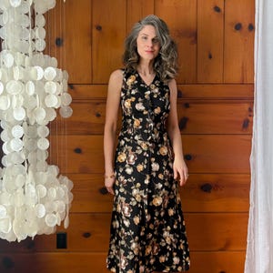 K&ouml;nnte beinhalten: Eine Frau tr&auml;gt ein schwarzes Midikleid mit Blumenmuster. Das Kleid hat einen V-Ausschnitt und eine Knopfleiste vorne. Sie tr&auml;gt braune Sandalen mit Absatz.