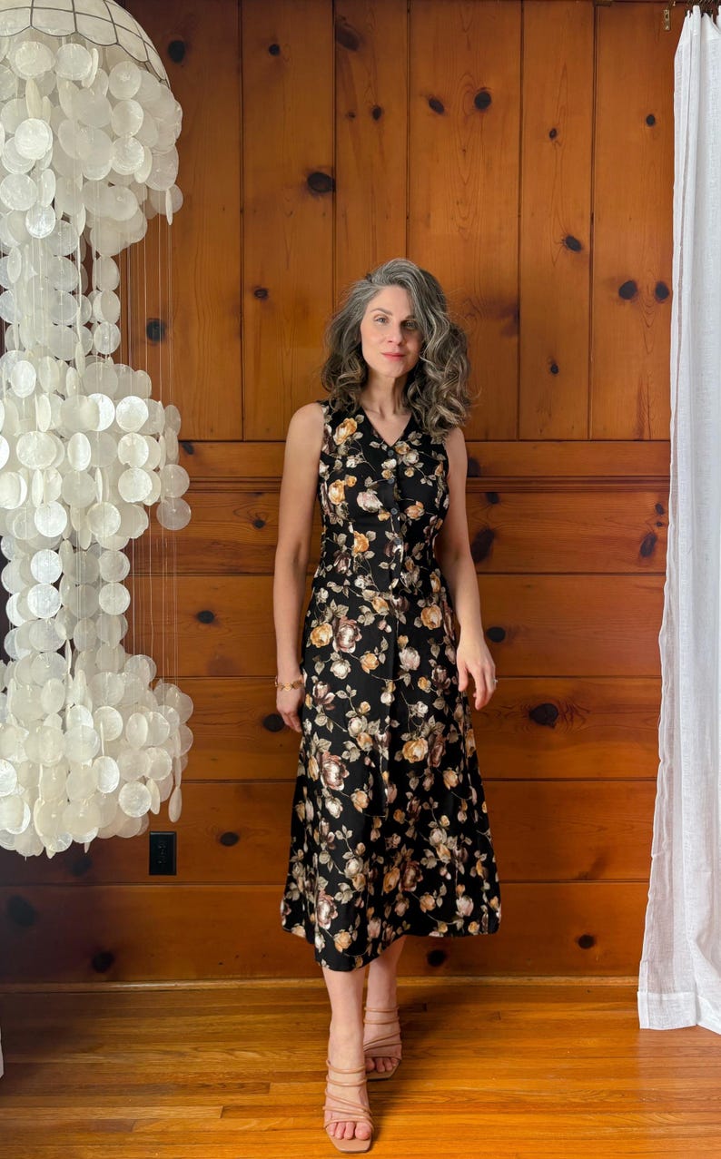 K&ouml;nnte beinhalten: Eine Frau tr&auml;gt ein schwarzes Midikleid mit Blumenmuster. Das Kleid hat einen V-Ausschnitt und eine Knopfleiste vorne. Sie tr&auml;gt braune Sandalen mit Absatz.