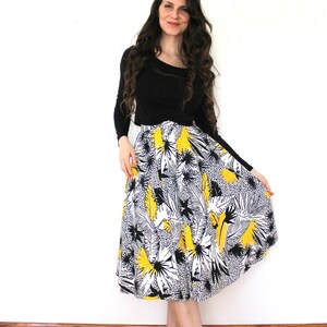 May include: A woman wearing a black long-sleeved shirt and a yellow, black, and white floral midi skirt. She is standing on a wooden floor with a white wall behind her.