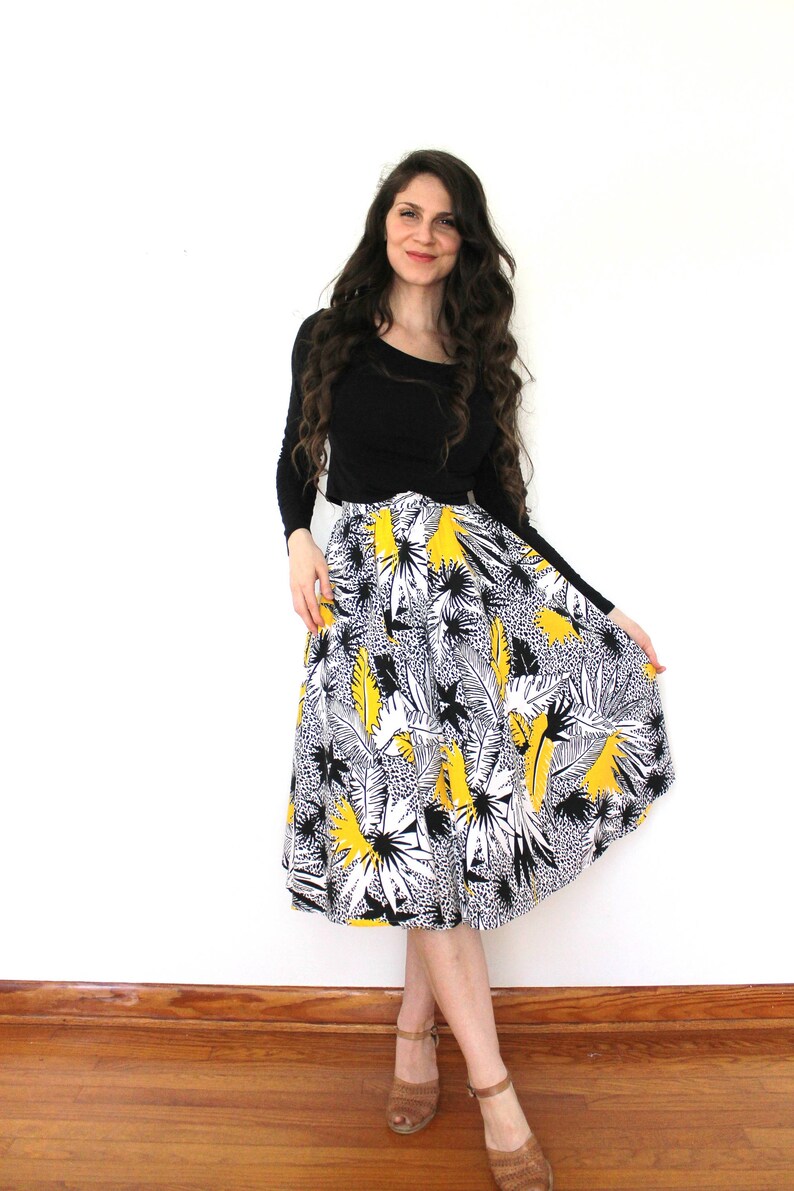 May include: A woman wearing a black long-sleeved shirt and a yellow, black, and white floral midi skirt. She is standing on a wooden floor with a white wall behind her.