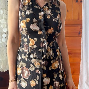 K&ouml;nnte beinhalten: Ein &auml;rmelloses schwarzes Kleid mit einem Blumendruck mit braunen, gelben und rosa Rosen. Das Kleid hat einen Knopfleisten vorne und ein tailliertes Oberteil.